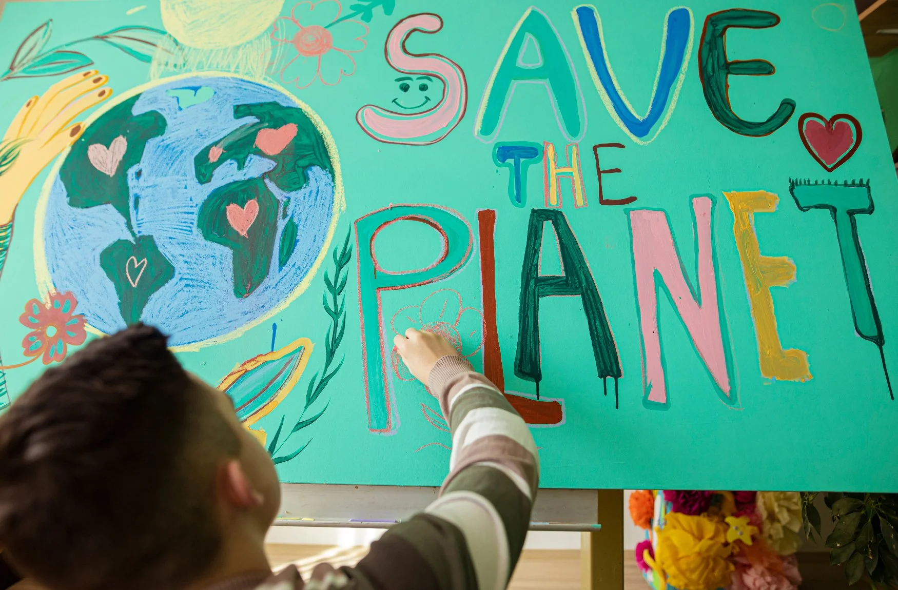 Person drawing a flower on a colorful poster with the text 'Save the Planet' and a drawing of Earth surrounded by hearts, flowers, and leaves.