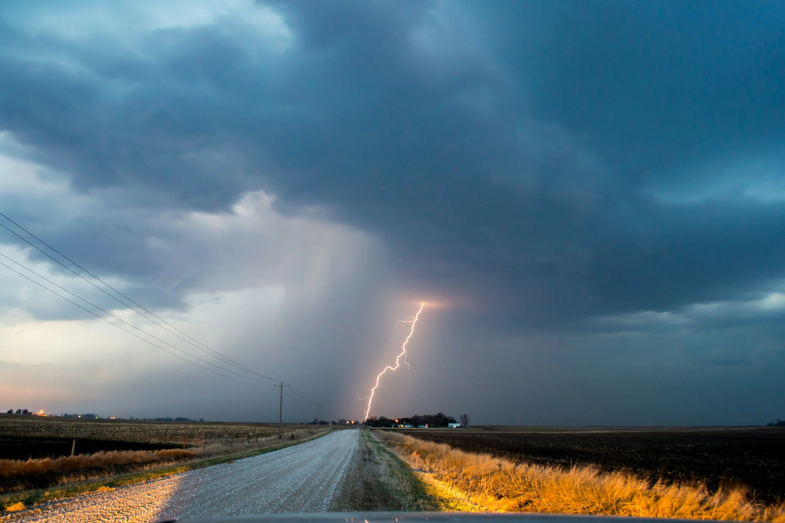 Lightning strikes in a stormy sky over a rural dirt road with fields on either side.