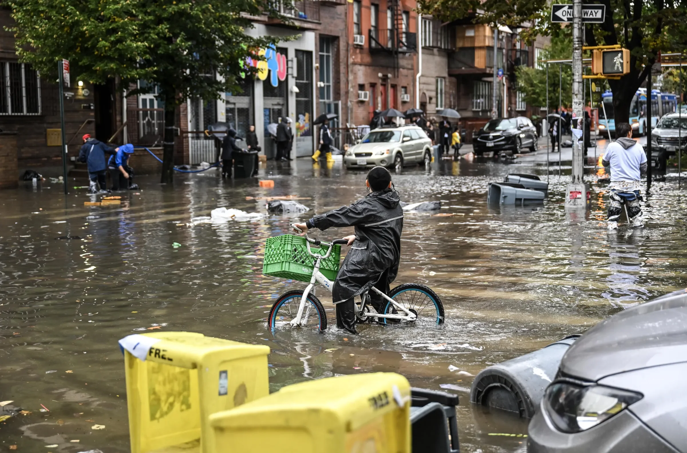 A flooded city street with people walking through the water, some with umbrellas and boots, cars partially submerged, and a person with a bicycle carrying a green crate in the foreground.