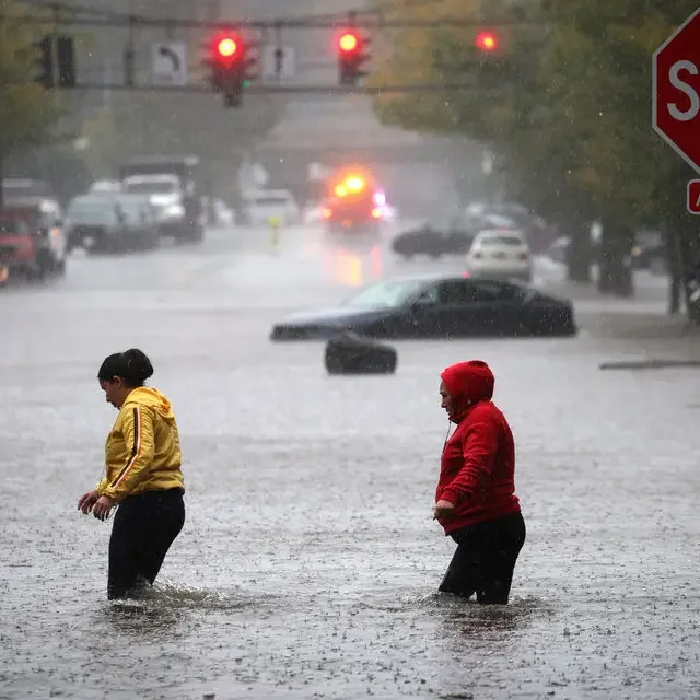 Two people wading through a flooded intersection in heavy rain, one in a yellow jacket and one in a red hooded jacket, with stalled cars, red traffic lights, and a stop sign visible.