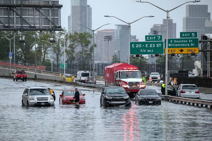 Flooded city street with submerged cars, emergency responders, and highway signs overhead.