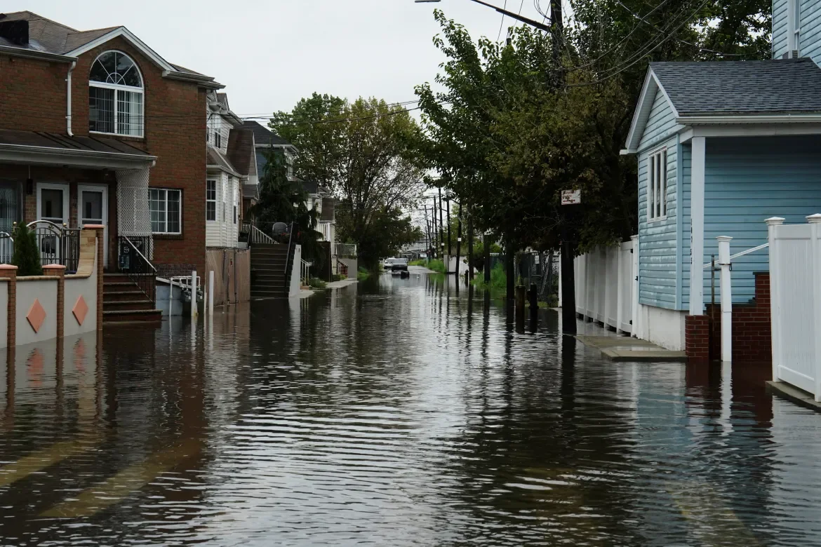 A flooded residential street with water covering the road and partially submerging the curb, houses on both sides, and trees in the background under an overcast sky.