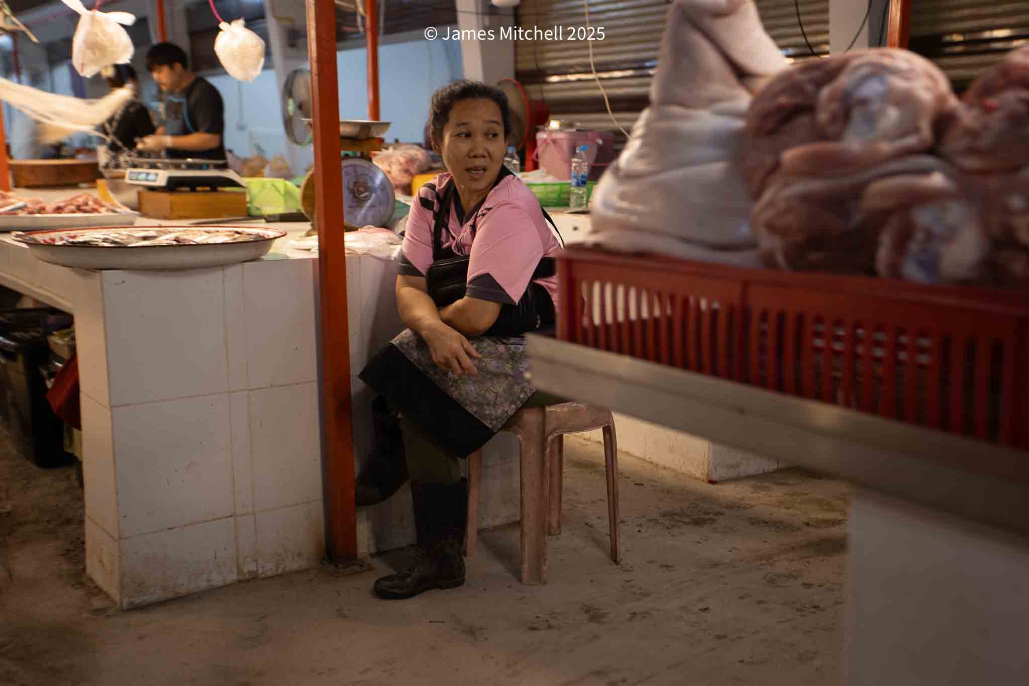A woman sitting on a small plastic stool behind a meat stall at a market, surrounded by various cuts of meat and a scale, with a worker in the background preparing meat.
