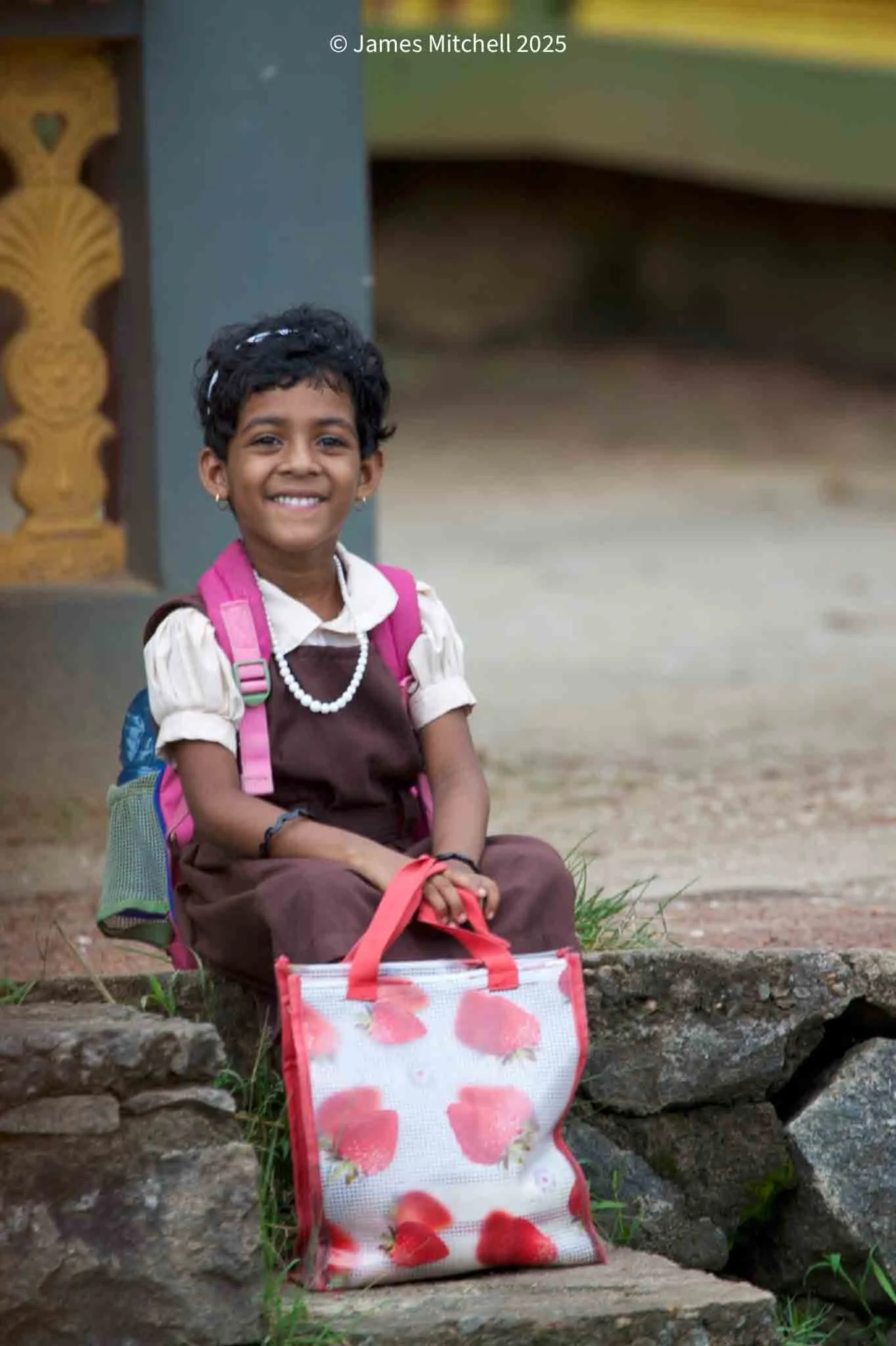 A smiling young girl with short curly hair, wearing a school uniform and jewelry, sitting on a stone ledge outside. She has a pink backpack and a floral tote bag with red flowers.