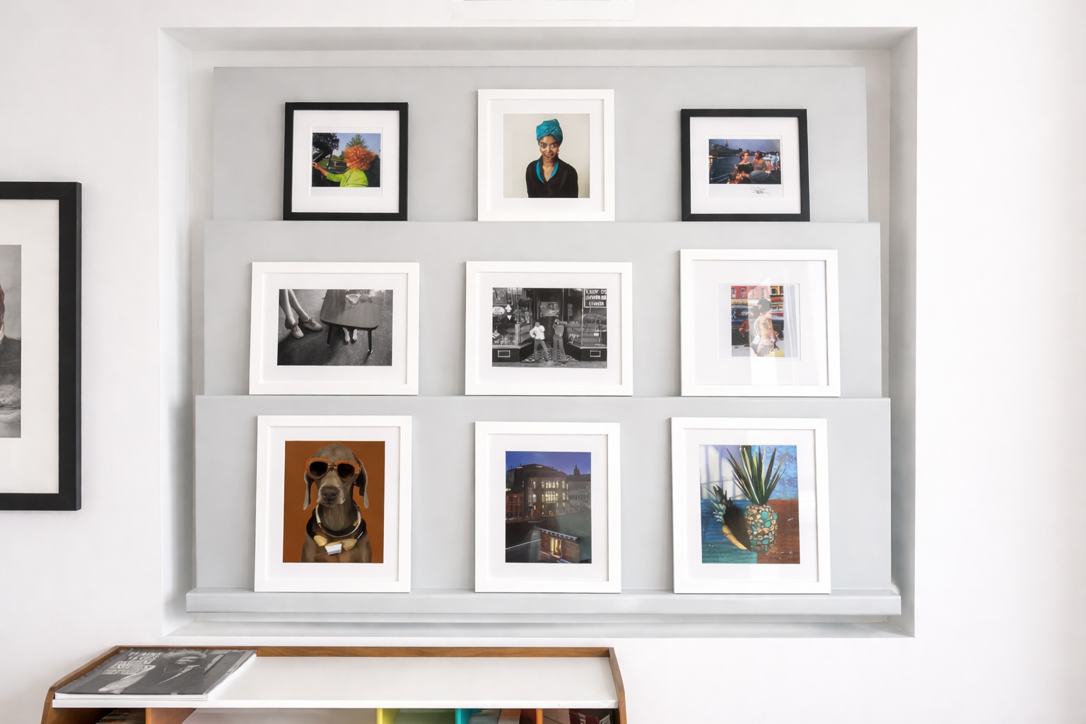 Wall display with nine framed artworks arranged in three rows on a white wall, including photographs of people and objects, a dog with sunglasses, and a painting of a plant.