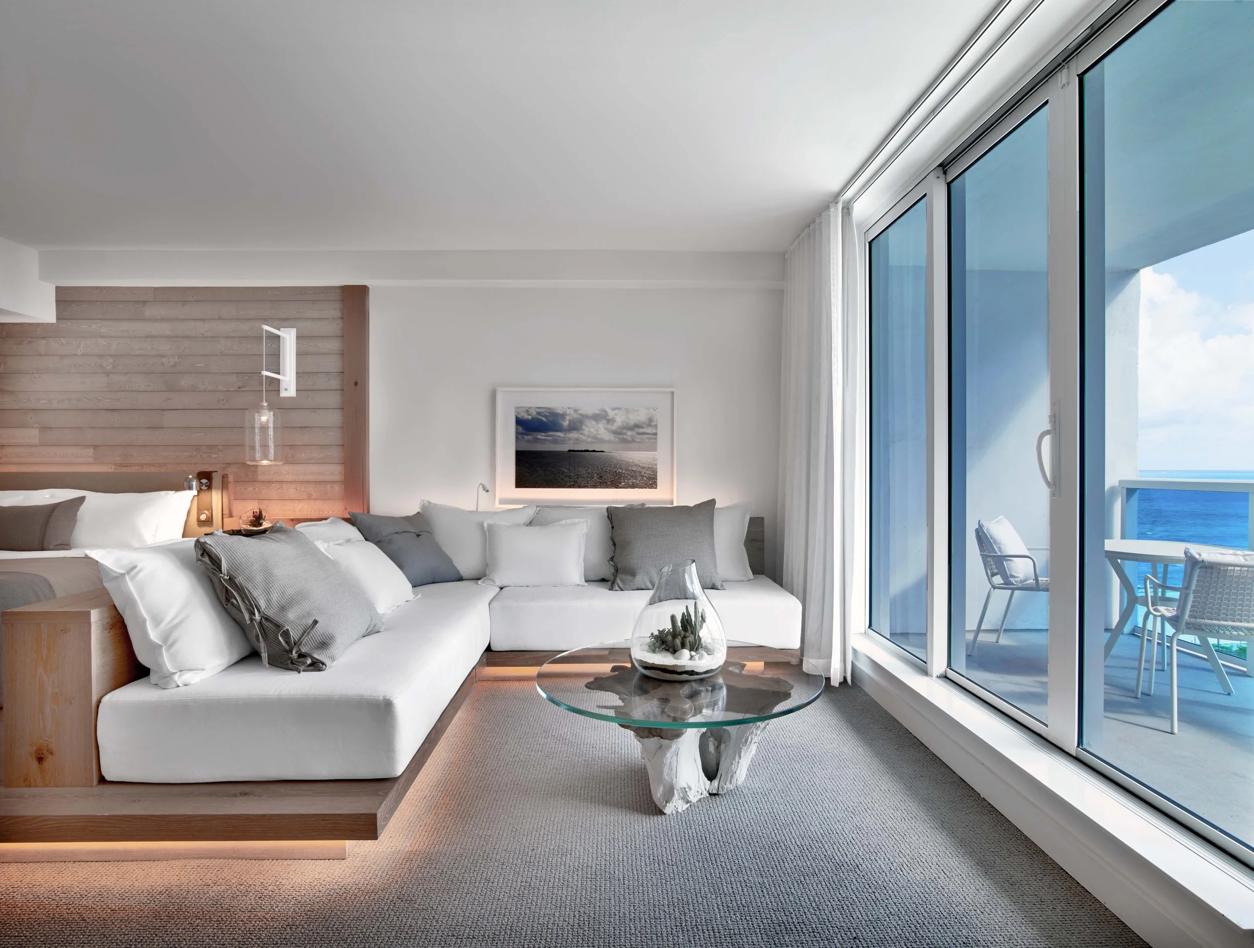 A living room with a large corner sofa with multiple white and gray cushions, a glass coffee table with a decorative centerpiece, large sliding glass doors opening to a balcony with a view of the ocean and sky, and a picture on the wall depicting a s