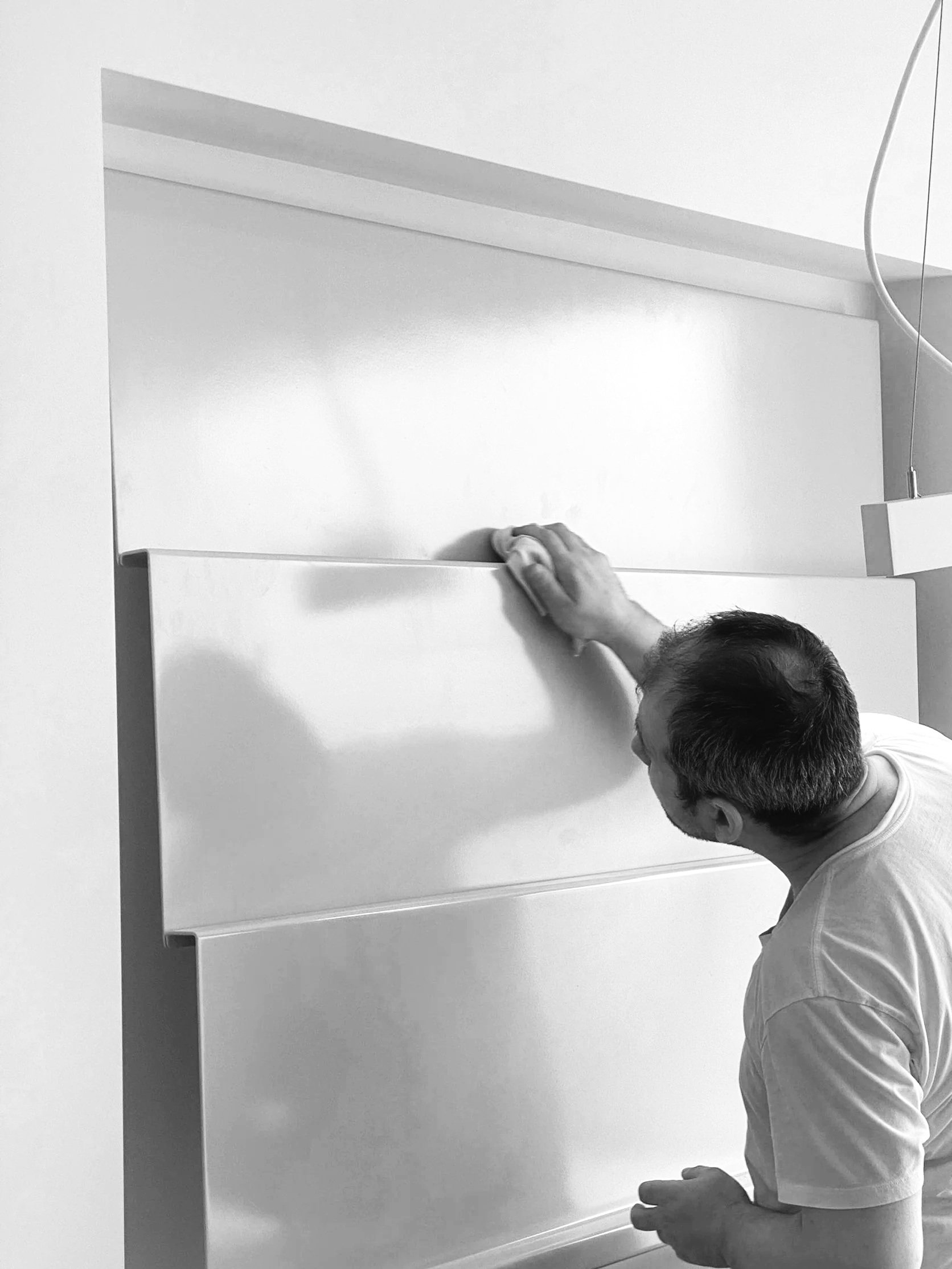 A person with short hair wearing a white shirt is fixing or assembling a white cabinet or shelf unit against a white wall.