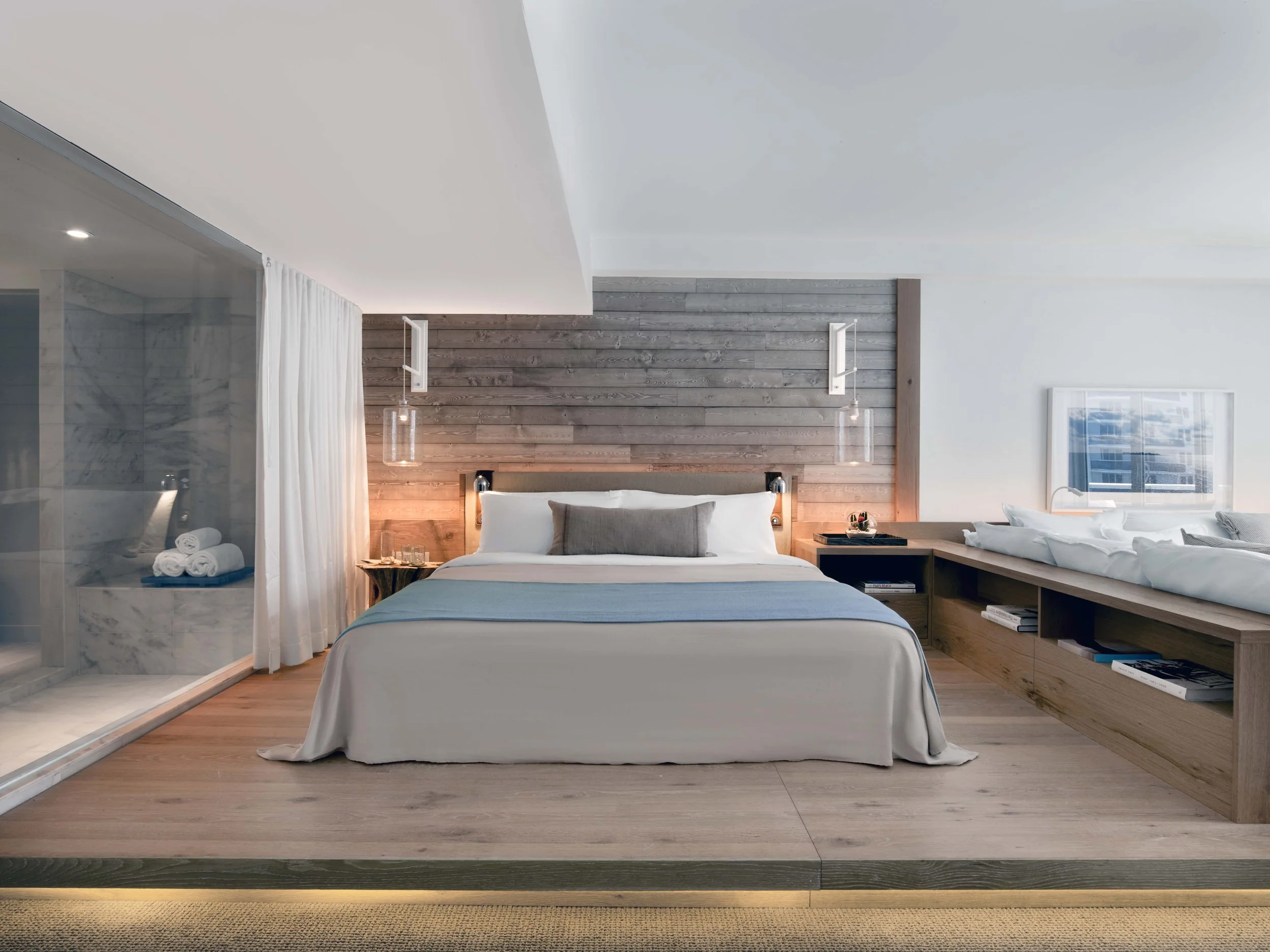Modern bedroom with a white bed, gray pillows, wooden headboard, and wooden accents; bathroom with marble shower, white towels, and glass enclosure; wood-paneled wall with hanging pendant lights; window showing cityscape.