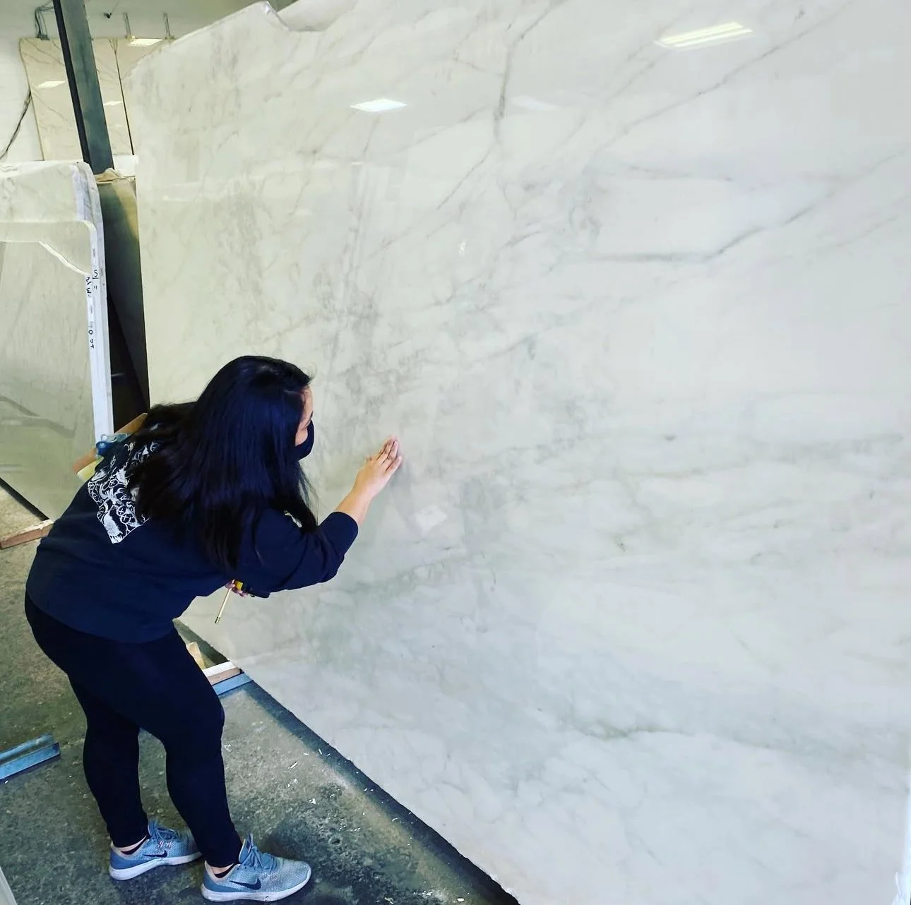 Woman wearing a black mask touching a large slab of white marble with gray veins in a warehouse or showroom.