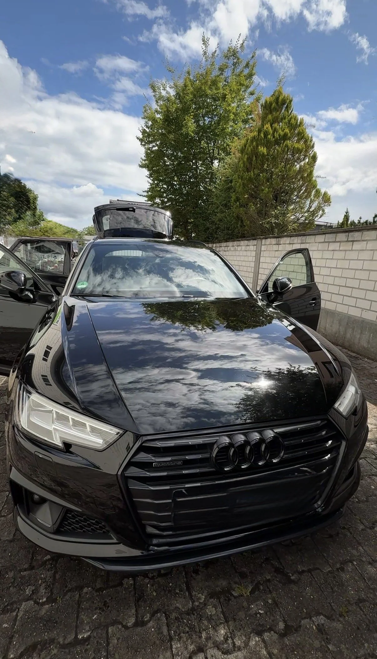 Schwarzer Audi-A4 Avant mit reflektierendem Lack, geöffneten Türen und Dachbox, parkend neben einer hohen Mauer, im Hintergrund Bäume und ein blauer Himmel mit Wolken. Lackpolitur FJayAutoCare
