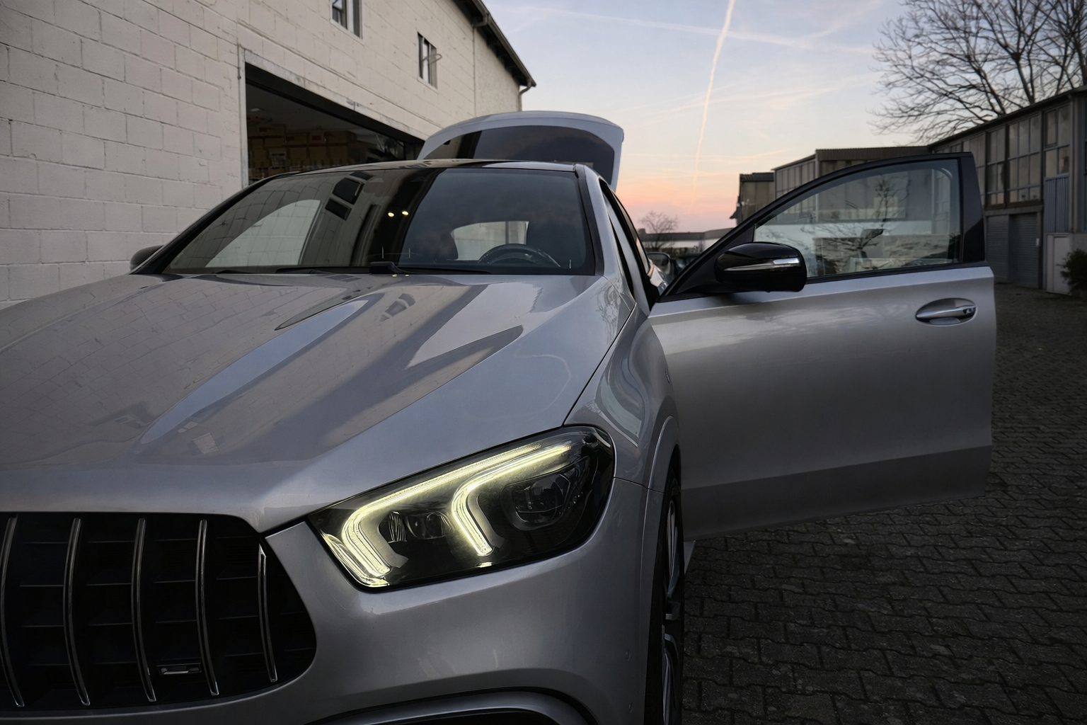 Silberner SUV vor einer Garage bei Sonnenuntergang, mit offenem Kofferraum und Fahrertür.