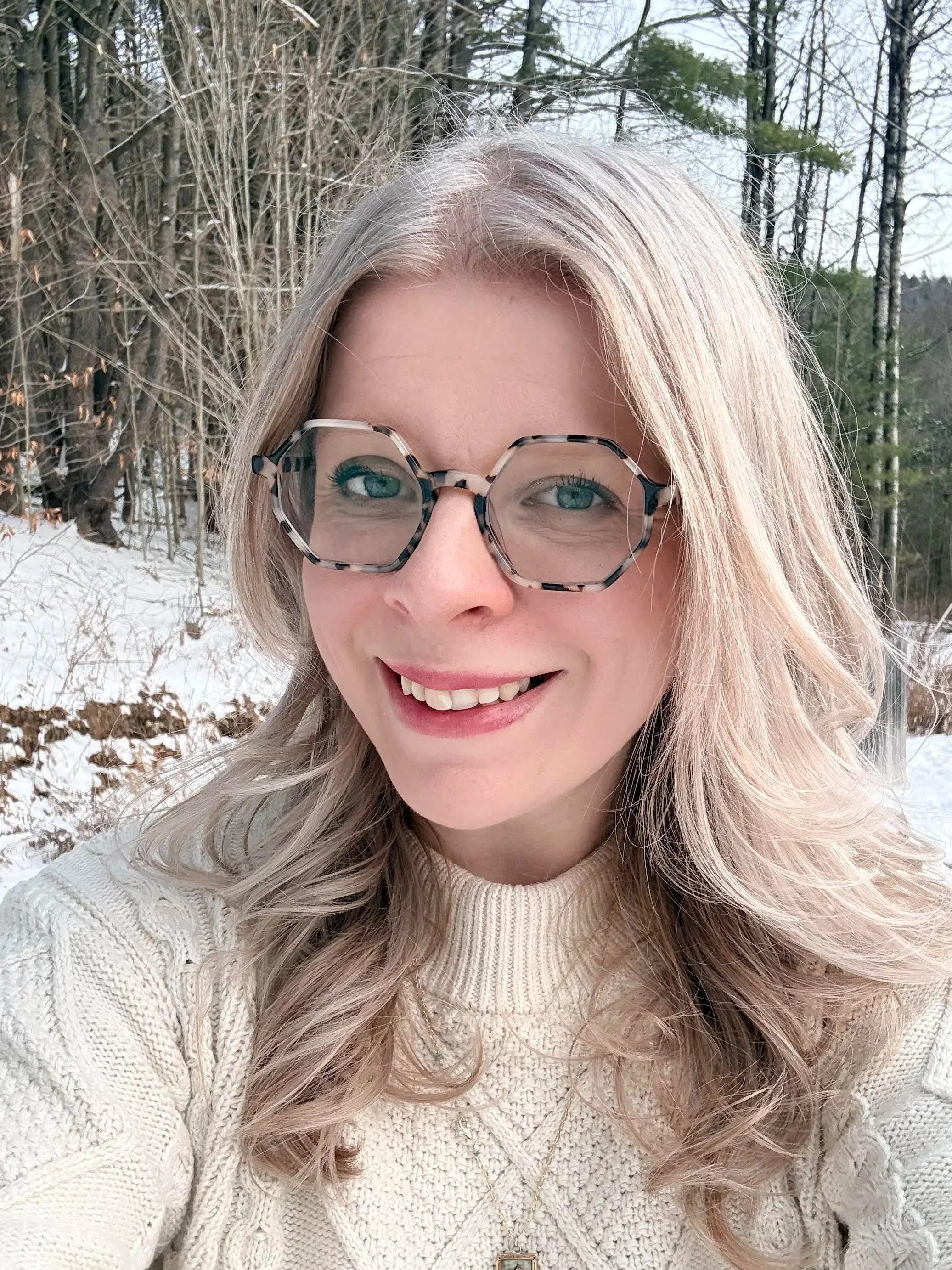 A woman with blonde hair and wearing large, patterned glasses smiles for a selfie outdoors in a snowy, wooded area.