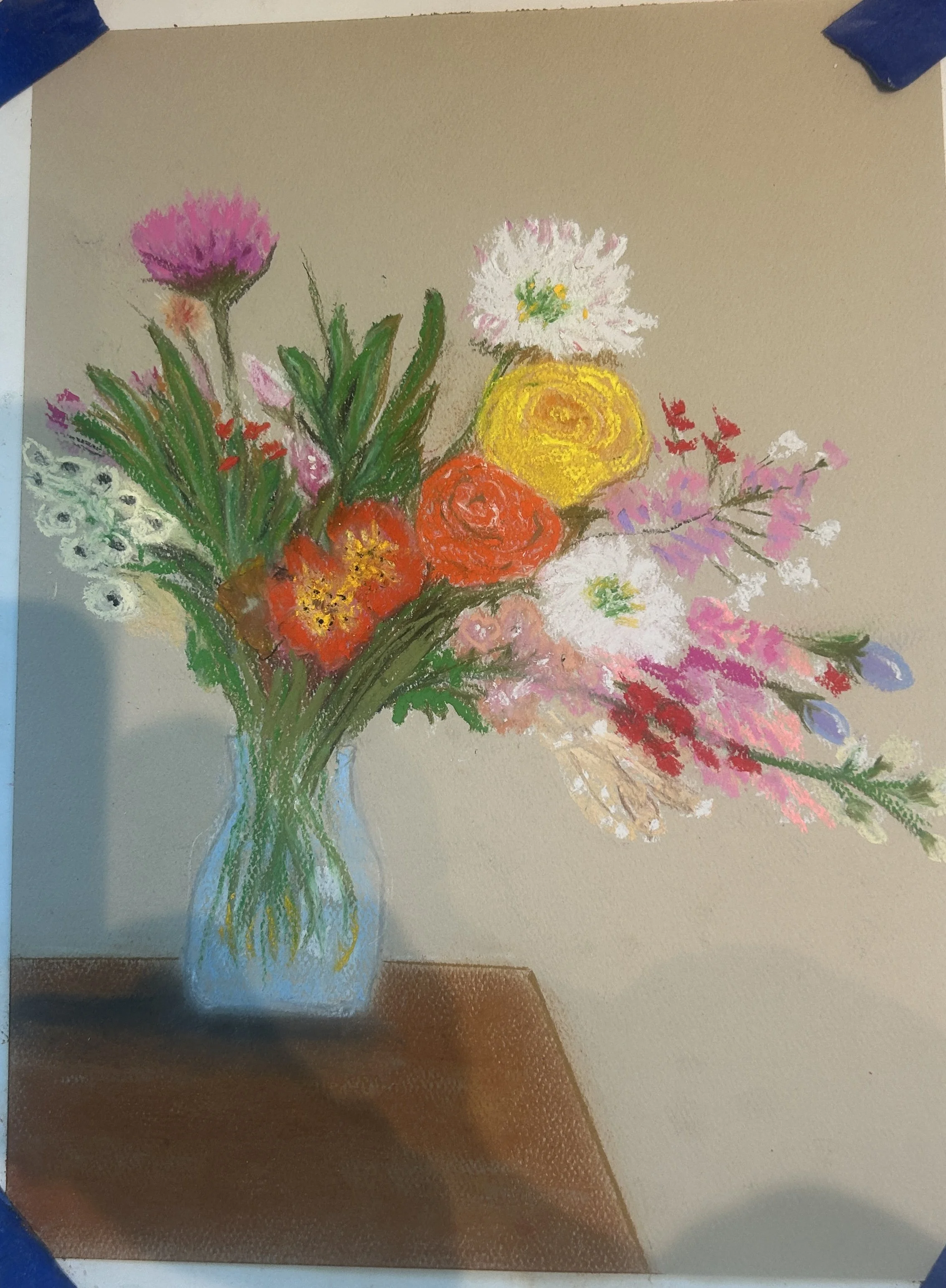 A colorful flower bouquet in a glass vase on a wooden surface, featuring pink, white, yellow, orange, and red flowers with green leaves.