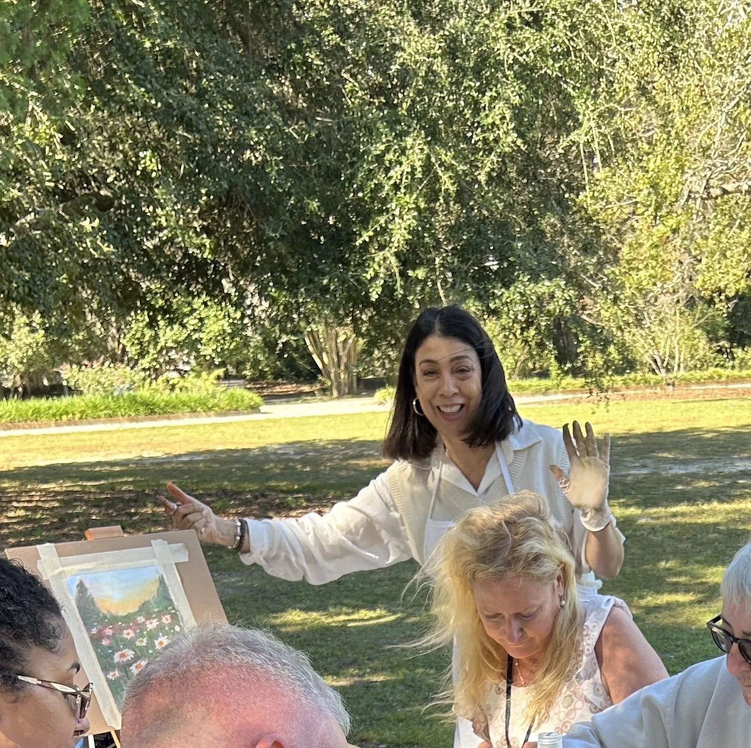 A woman with dark hair, smiling and waving, standing outdoors under a large leafy tree, with a group of people sitting at a table, some looking at artwork on the table, in a park setting.