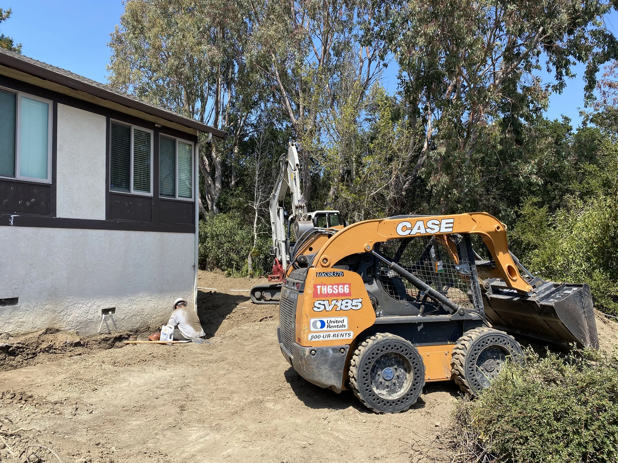 A construction worker kneeling on the ground near the foundation of a house, digging or working with tools. A small, orange CASE skid-steer loader is parked nearby on a dirt site, with a backdrop of trees and a clear blue sky.