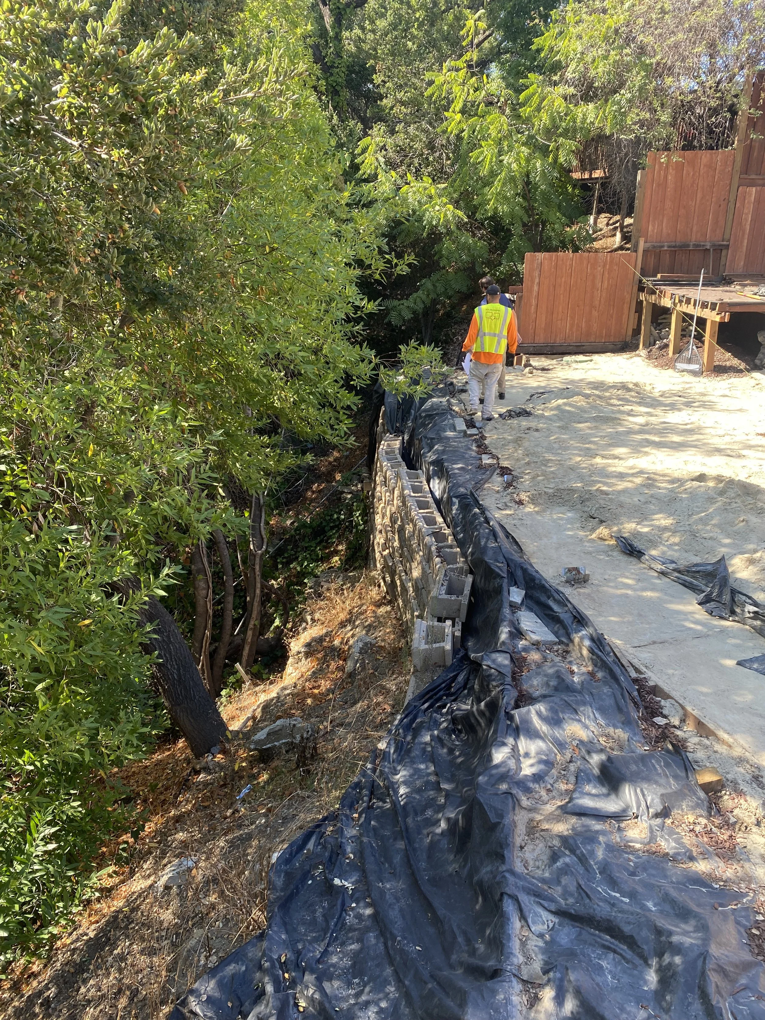 Construction workers in safety vests and helmets working along a hillside with a temporary wooden fence, black plastic sheeting, and dirt at a construction site.