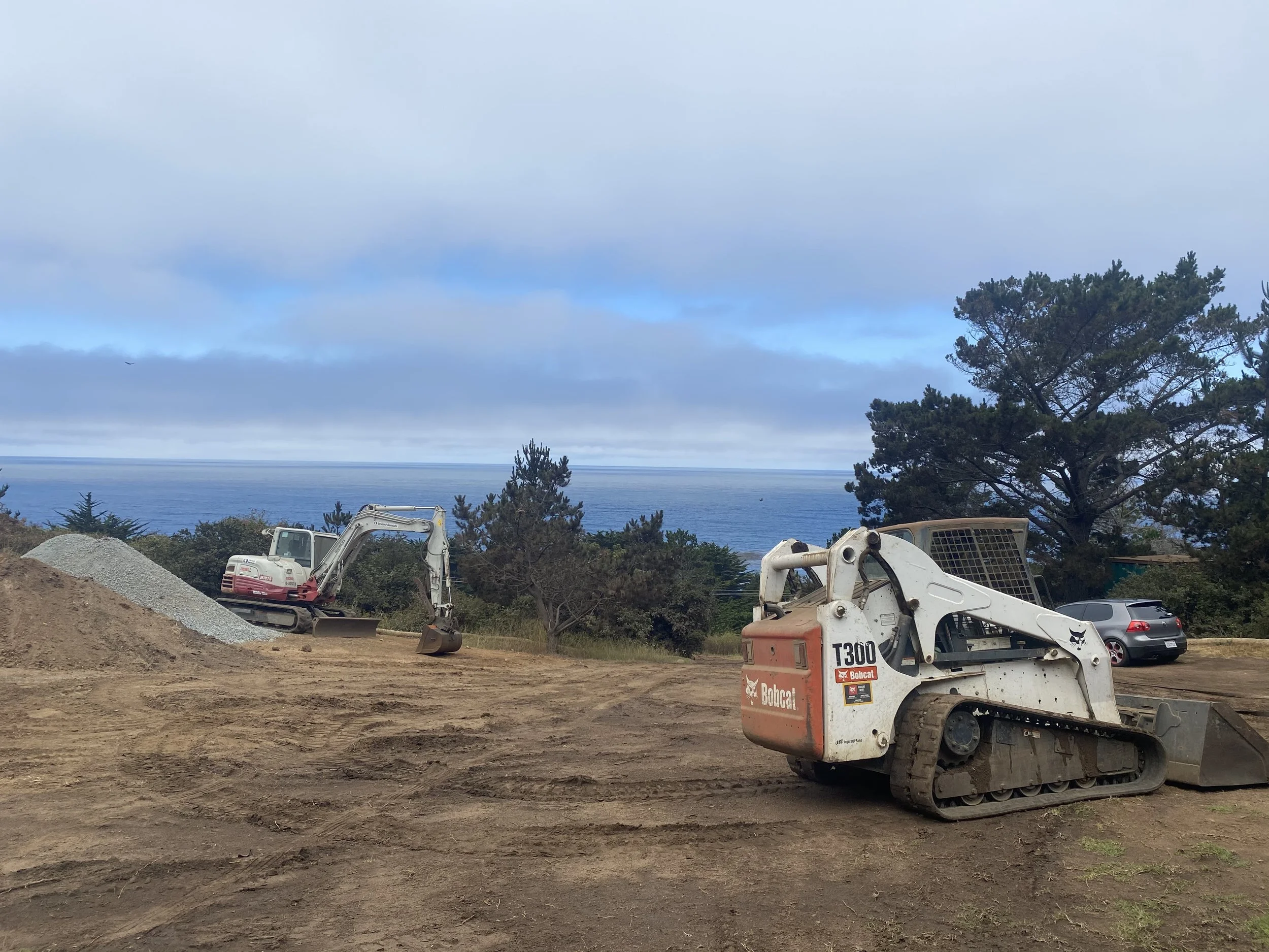 Construction site with two compact excavators and a pile of gravel, overlooking an ocean view with trees and cars parked nearby.