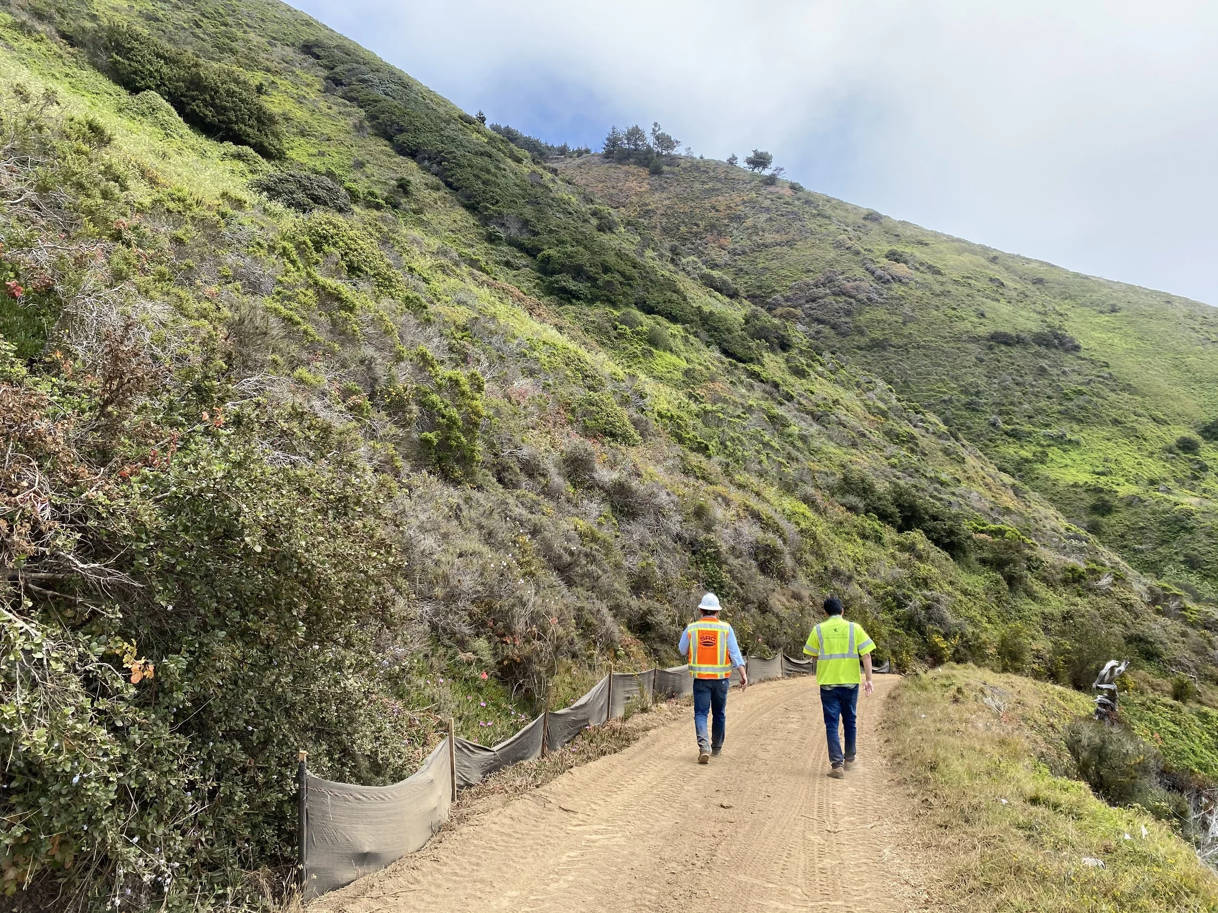Two construction workers walking along a dirt path on a hillside, with lush green vegetation and a partly cloudy sky.