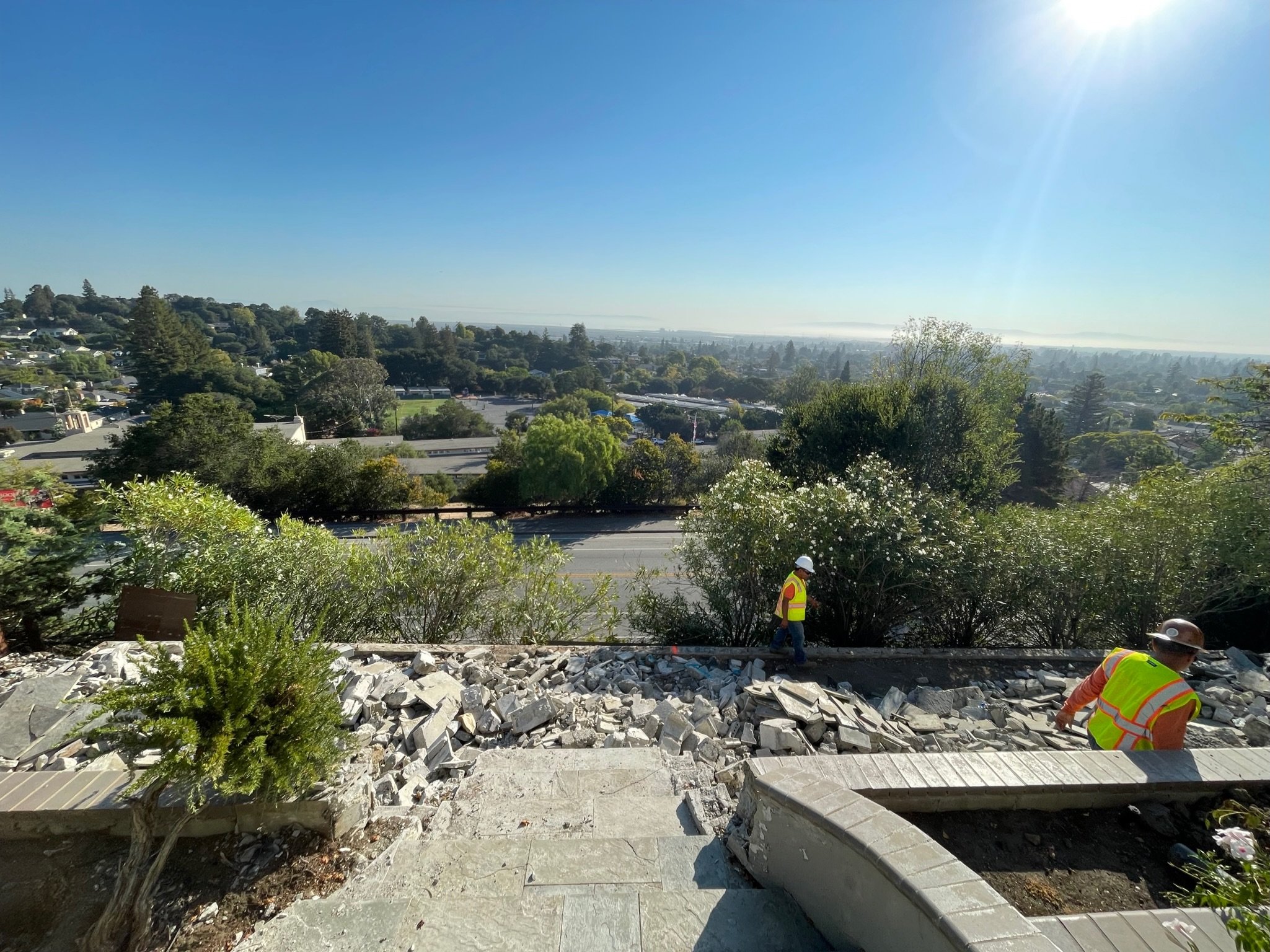 Construction workers in safety vests and helmets working on a sidewalk renovation on a sunny day, overlooking a cityscape with trees and buildings in the distance.
