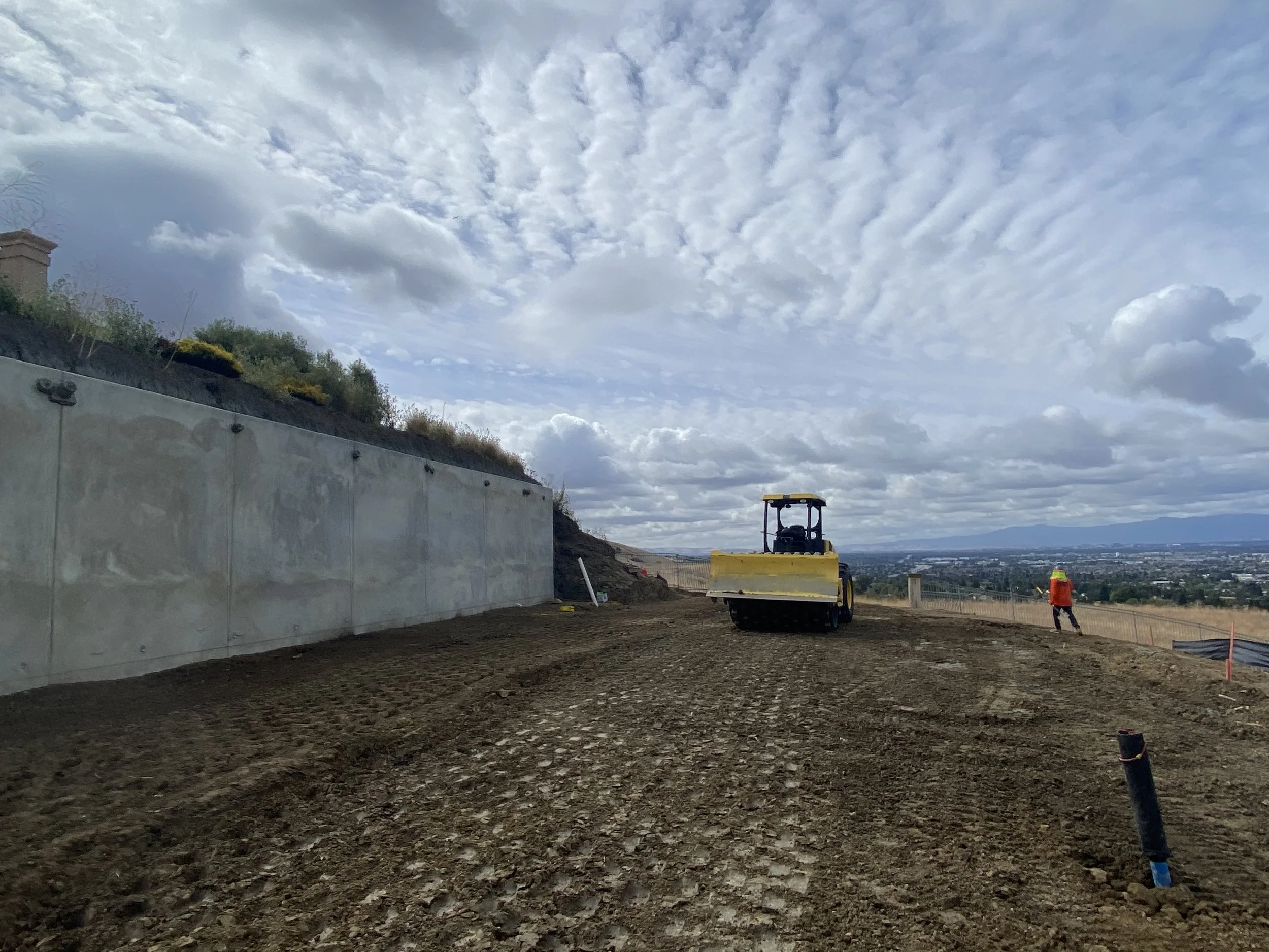 A construction site on a hillside with dirt ground, a yellow ride-on roller compactor, a worker in an orange safety vest and helmet, and a concrete retaining wall on the left, with a view of a cityscape and cloudy sky in the background.