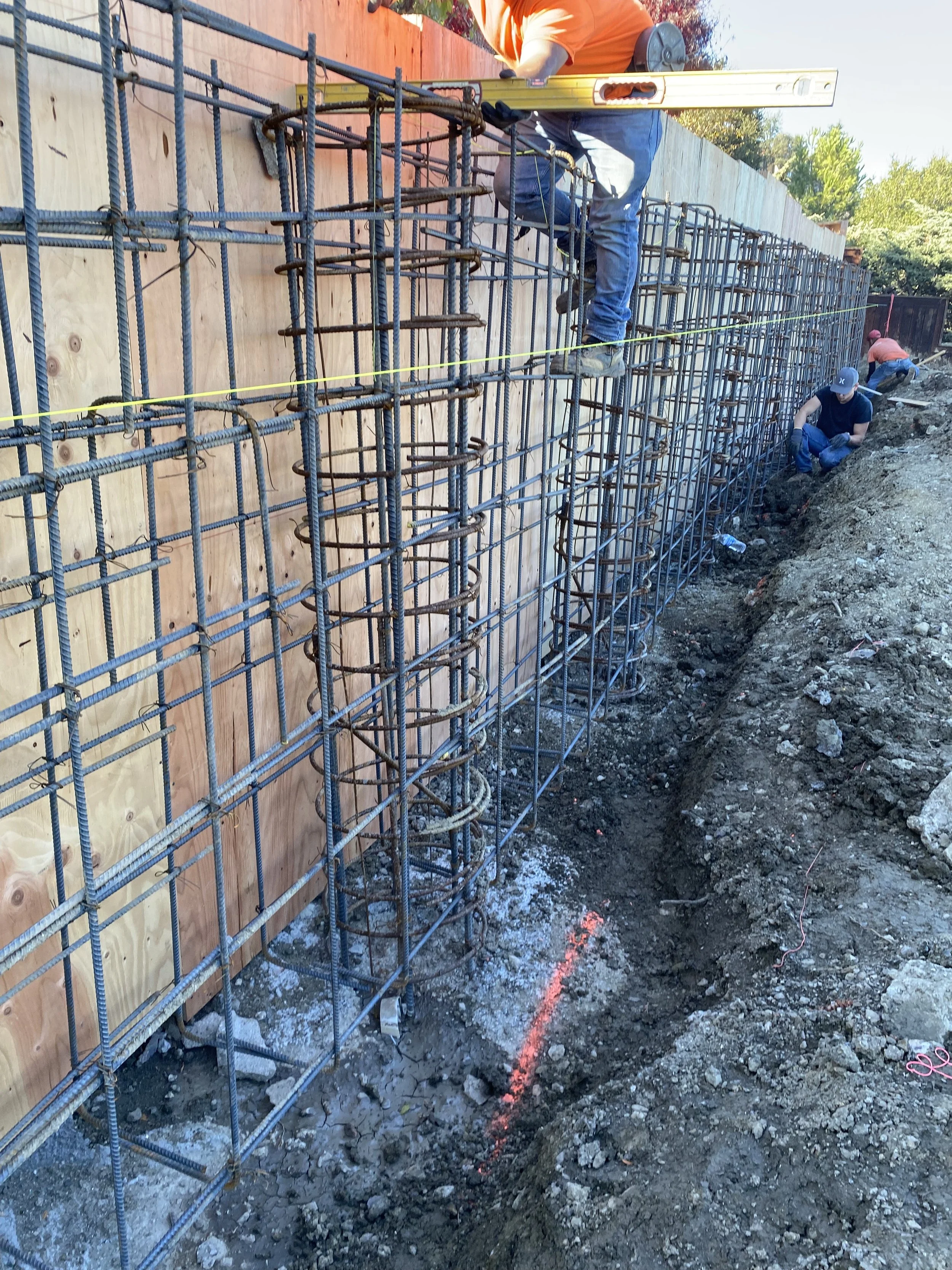 Workers assembling reinforced steel rebar framework on a construction site with wooden formwork, black soil, and a yellow level tool.