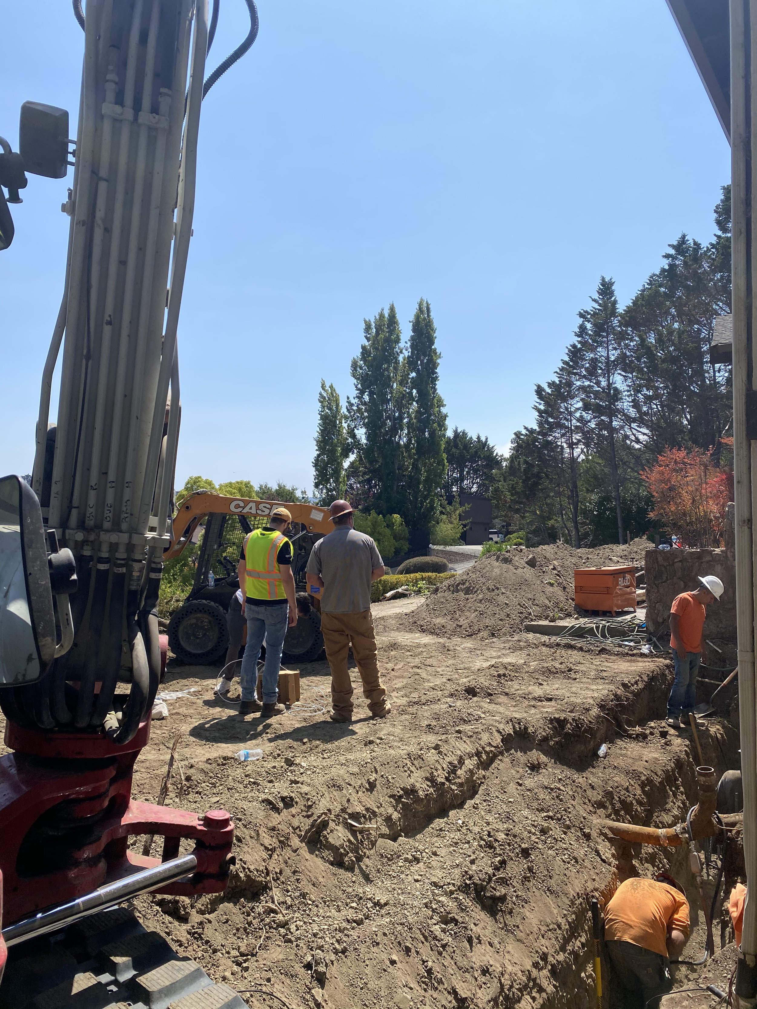Construction workers on a site working with excavated soil, machinery, and underground pipes, with trees and a house in the background.