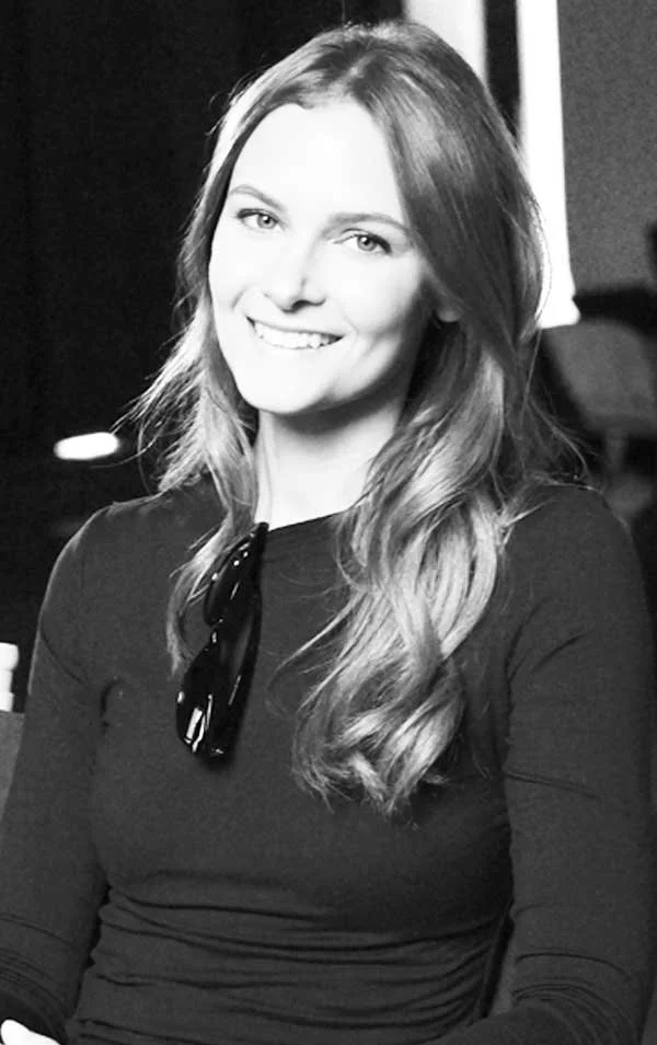A young woman with long wavy hair wearing a black top and sunglasses hanging around her neck, smiling for the camera in a black and white photo.