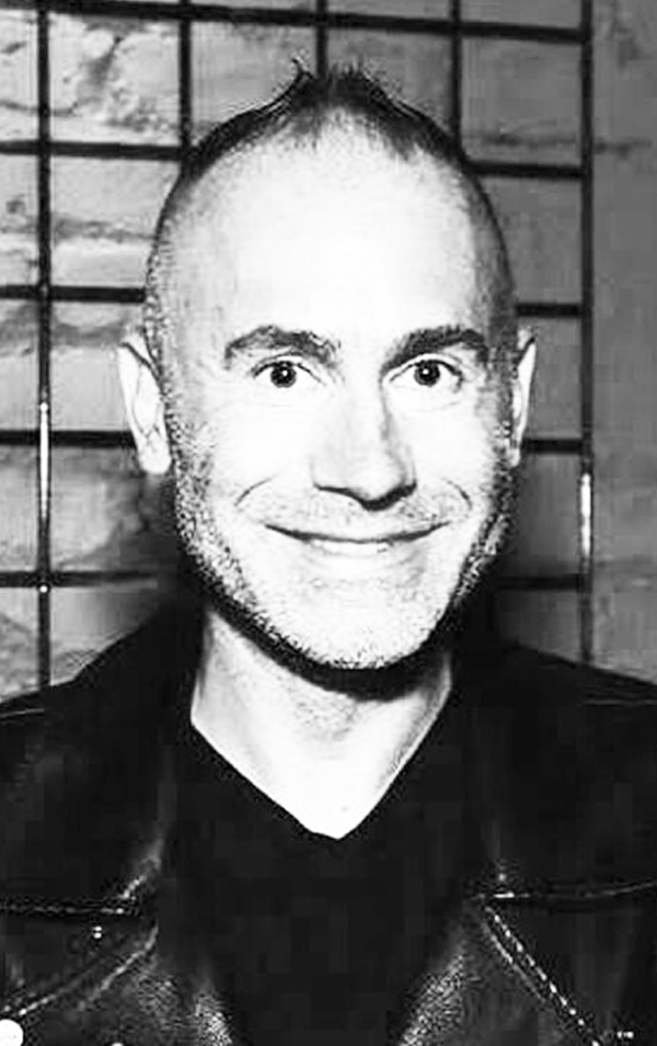 Black and white photo of a smiling man with short hair and a beard, wearing a leather jacket. Background appears to be a tiled wall.