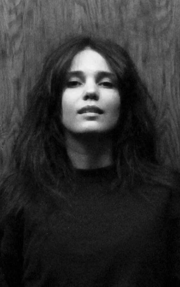 A black and white photo of a woman with messy hair, looking directly at the camera, standing against a wood-paneled wall.