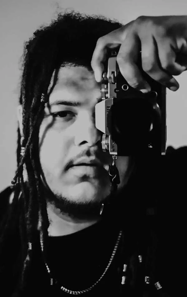 A person with dreadlocks holding a camera up to their face, taking a selfie in black and white.