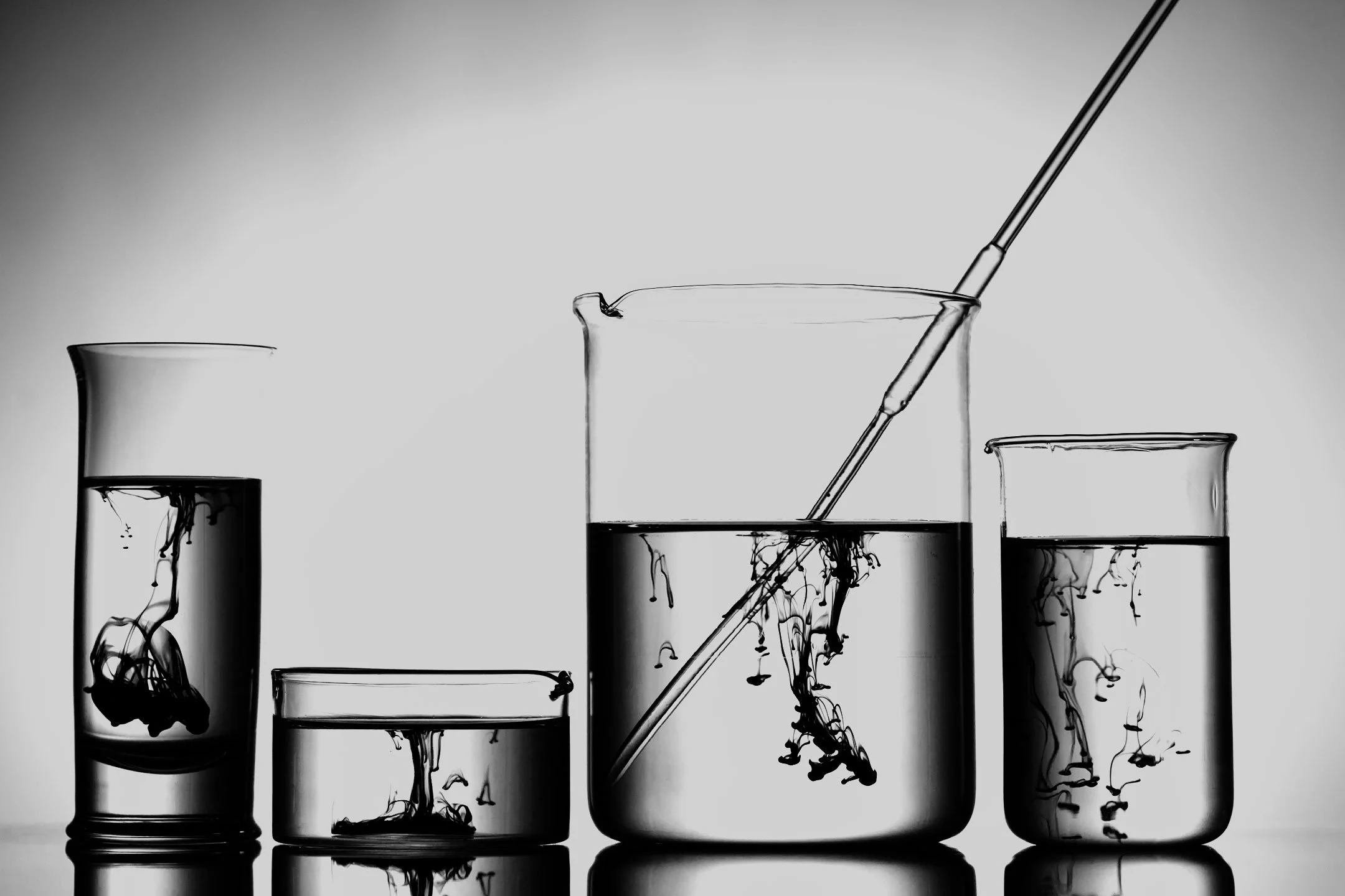 Black and white photo of four glass test tubes and a beaker filled with liquid, with one test tube containing a pipette.