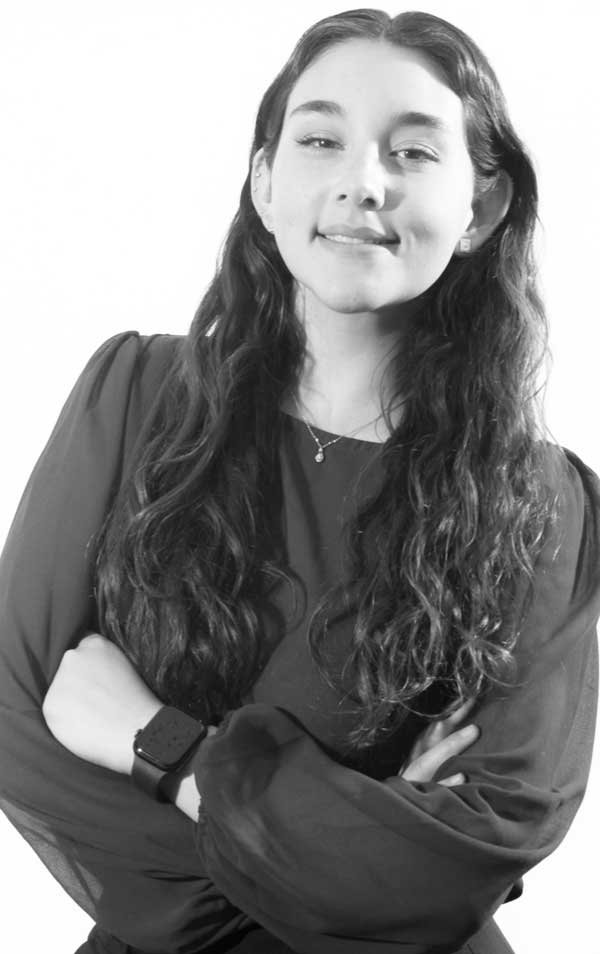 A young woman with long wavy hair, smiling and crossing her arms, wearing a dark blouse and a smartwatch, in a black-and-white portrait.