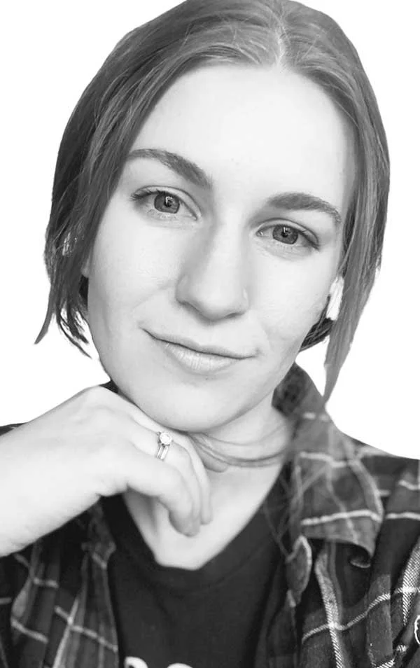 Black and white portrait of a young woman with short hair, wearing a checkered shirt and a ring on her finger, smiling gently.