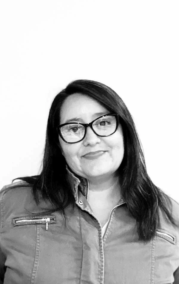 Black and white photograph of a woman with shoulder-length dark hair wearing glasses and a jacket, smiling at the camera against a plain background.