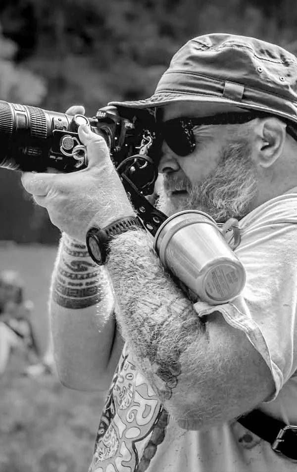 A man with a hat, sunglasses, and a beard is aiming a camera with a long lens. He has tattoos on his arms and is wearing a watch. A water bottle is attached to his arm. The background is blurred.