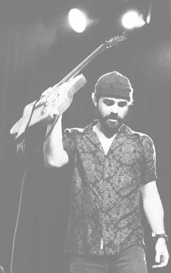 A man with a beard and mustache, wearing a patterned shirt and a headband, holding a guitar over his shoulder, standing on stage with bright lighting.