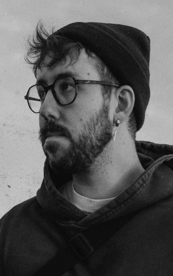 A young man with glasses, a beard, and a mustache wearing a beanie, earrings, and a hoodie, looking to the side in a black-and-white photo.