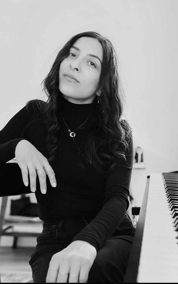 A woman with long wavy dark hair wearing a black turtleneck and jewelry, sitting at a piano with her right arm resting on the instrument in black-and-white. She is looking at the camera with a slight smile.