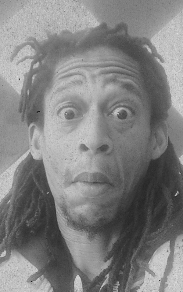 A black-and-white selfie of a man with dreadlocks lying on the floor, looking up at the camera with wide eyes and a surprised expression.