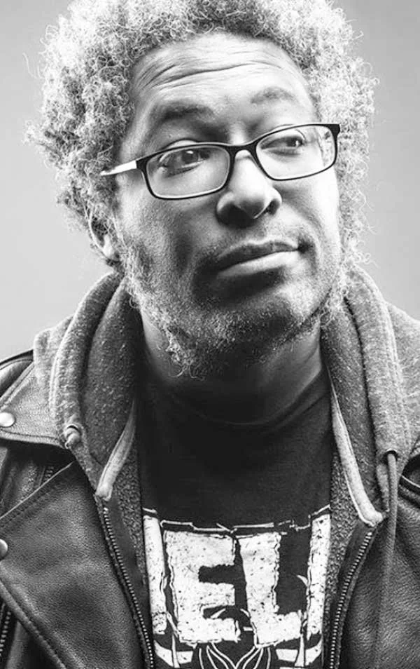 Black and white photo of a man with curly hair wearing glasses, a leather jacket, and a graphic t-shirt.
