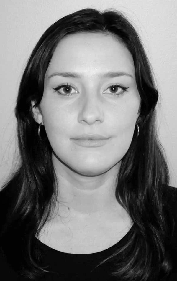 Black and white portrait of a young woman with long dark hair, wearing hoop earrings and a dark top, looking directly at the camera with a neutral expression.