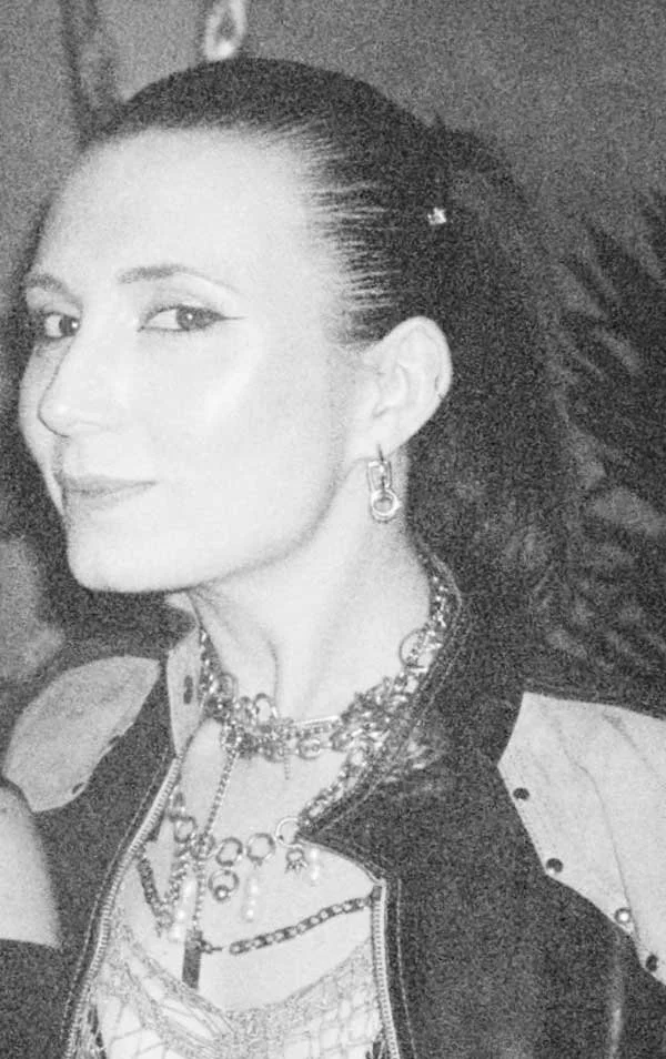 A woman with dark hair tied back, smiling slightly, wearing earrings, layered necklaces, and a jacket with shoulder patches, in a black-and-white photo.