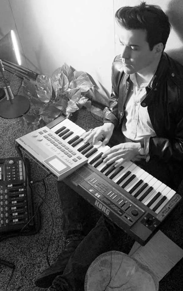A man playing a Korg keyboard synthesizer in a music studio with a small electronic drum pad and mixing console nearby.