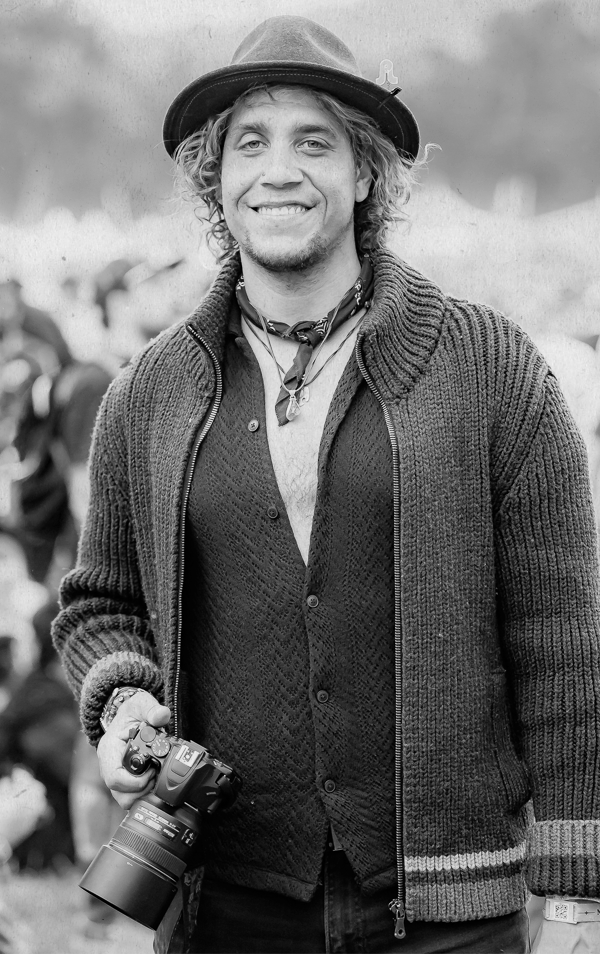 Black and white photo of a smiling man wearing a hat and a knitted sweater, holding a professional camera in his right hand, standing outdoors with blurred people and landscape in the background.