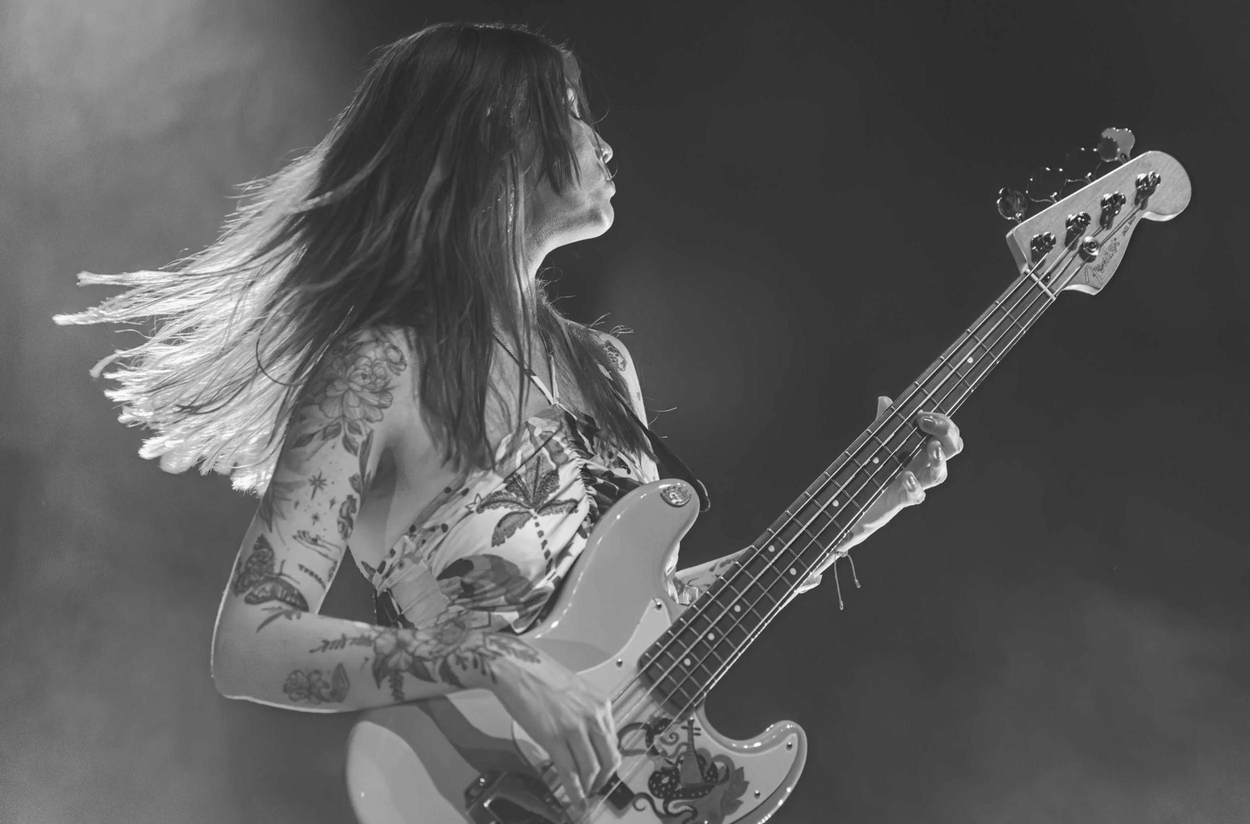 A female musician with tattoos playing an electric bass guitar on stage with her hair moving, captured in black and white photo.