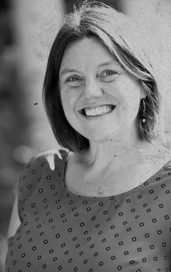 Black and white portrait of a smiling woman with shoulder-length hair, wearing a patterned blouse, standing outdoors near a concrete wall.