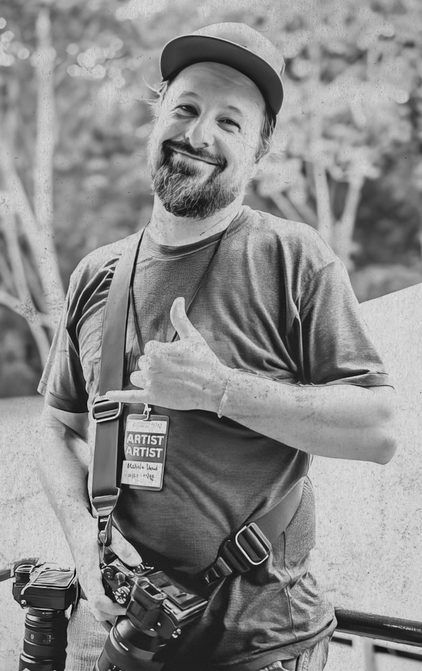 A smiling man with a beard wearing a baseball cap, artist badge, and camera gear, giving a thumbs up outdoors.