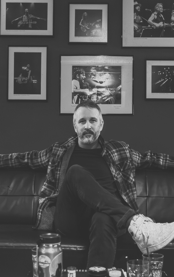 A man with a beard and short hair sitting on a black leather couch with his arms resting on the back, wearing a plaid shirt and black pants, in a room with framed band photos on the wall behind him.