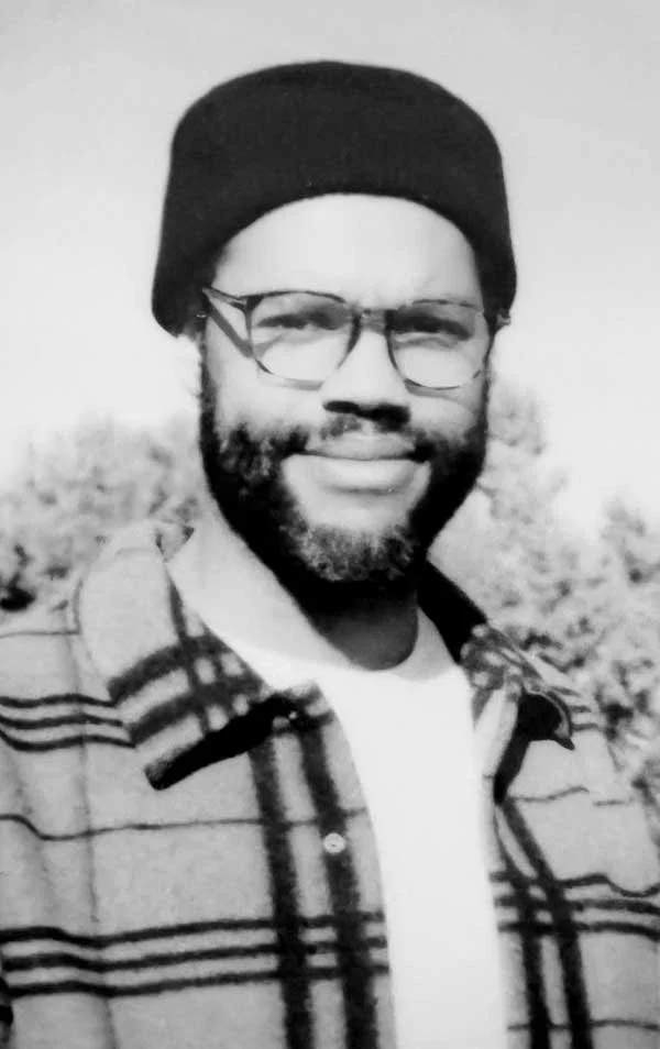 Black and white photo of a man with a beard, glasses, wearing a beanie hat and a plaid jacket, smiling outdoors.