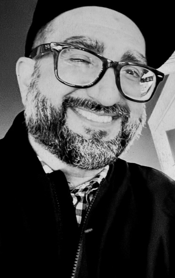A smiling man with glasses, a beard, and a beanie, wearing a checkered shirt and a black jacket, in black and white.